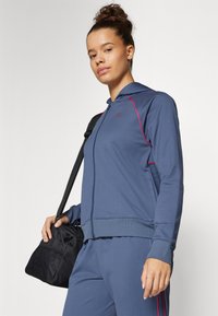 Blå idretts zip-up hettegenser med rød kantbånd, med front glidelås, hette og ribbestrikkede mansjetter. Kombinert med en svart veske.