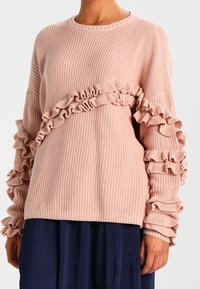 Różowy sweter z dzianiny z falbankami wzdłuż rękawów. Posiada okrągły dekolt i fakturowaną tkaninę, zestawiony z plisowaną ciemną spódnicą.