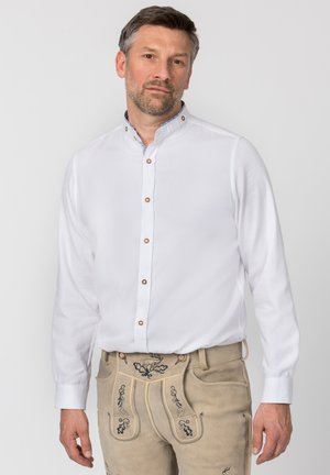 Man wearing white buttoned shirt with mandarin collar and beige leather pants with embroidered floral designs, standing against plain background.