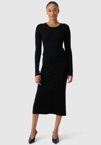 Zwarte geribbelde lange mouw top gedragen met een zwarte stretchy midi rok. Gladde textuur en aansluitend ontwerp, compleet met puntige zwarte hakken.