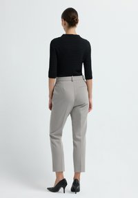 Woman with hair in bun wearing black textured sweater, gray high-waist pants, and black woven high heels, standing facing away on white background.