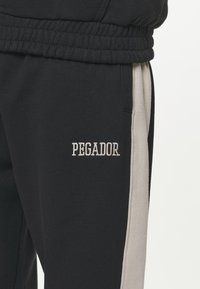 Pantalones de chándal negros con franjas laterales beige, con cintura elástica y bolsillos. La palabra "PEGADOR" está bordada en blanco en la parte superior del muslo.