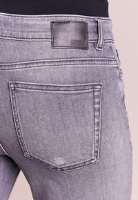 Vaqueros desgastados en gris con un parche de cuero etiquetado como "DRYKORN" en la parte trasera, con bolsillos estándar y detalles de costura visibles.