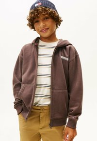 Sudadera marrón con cremallera y un logo, combinada con una camiseta de rayas y pantalones beige. La sudadera tiene capucha y bolsillos frontales.