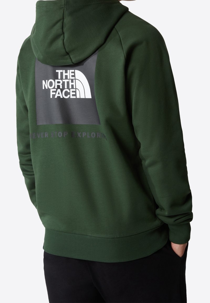 Personne portant un sweat à capuche vert foncé avec le logo "The North Face" et le texte partiellement visible "Never Stop Exploring" dans le dos.