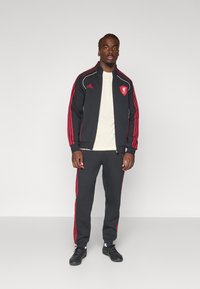 Zwart atletisch trainingspak met rode strepen, met een logo op de jas. Het geheel heeft een rits aan de voorkant en is gecombineerd met zwarte sneakers.