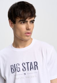 Camiseta de algodón blanca con un logo y texto desgastados en azul oscuro "BIG STAR" debajo. Manga corta y escote redondo.
