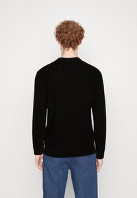 Schwarzer Strickpullover mit Rundhalsausschnitt und langen Ärmeln. Der Rücken hat einen gerippten unteren Saum und eine strukturierte Stoffoberfläche. Getragen mit blauen Jeans.