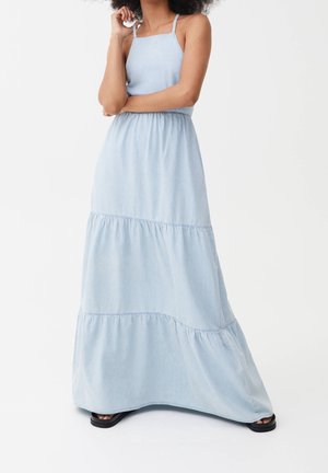 Femme portant une robe maxi à volants sans manches, de couleur bleu clair, avec des fines bretelles et des sandales noires, debout devant un fond blanc.