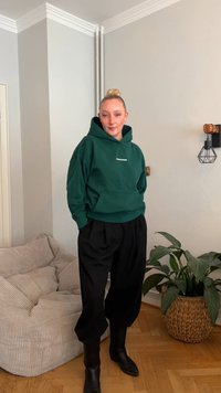 Sweatshirt verde com capuz, bolso frontal e texto branco, combinado com calças pretas largas e botas pretas de couro até ao tornozelo. Sofá bege texturizado e planta ao fundo.