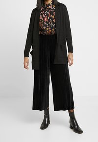 Cardigan negro, camisa floral y pantalones anchos de pierna plizada negros. El cardigan tiene un corte relajado y bolsillos; las botas son negras con punta cuadrada.