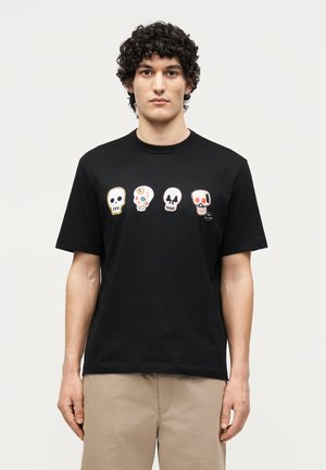 Άνδρας με σγουρά μαλλιά που φοράει μαύρο μπλουζάκι με τέσσερα πολύχρωμα σχέδια κρανίων στο στήθος και μπεζ παντελόνι, στέκεται μπροστά σε απλό φόντο.