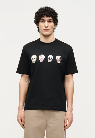 Hombre con pelo rizado que lleva una camiseta negra con cuatro diseños de calaveras coloridas en el pecho y pantalones beige, de pie frente a un fondo liso.