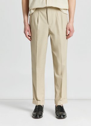 Persona con pantaloni beige plissettati con orli risvoltati e mocassini in pelle nera, in piedi davanti a uno sfondo chiaro e uniforme.