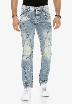 Verblasste hellblaue Jeans mit auffälligem Patchwork-Design, fünf Taschen und kontrastierenden Nähten. Kombiniert mit weißen Turnschuhen.