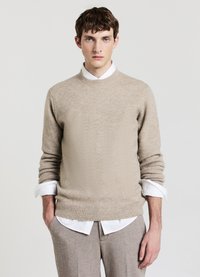 Beiger Strickpullover mit rundem Ausschnitt und geripptem Saum, getragen über einem weißen Hemd mit Kragen. Lange Ärmel mit aufgekrempelten Manschetten.