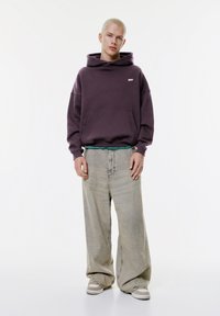 Sweat à capuche violet surdimensionné avec poche avant et logo, associé à un jean ample gris clair et des baskets grises avec des accents blancs.