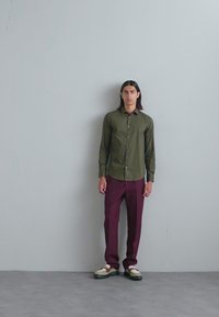 Olivengrøn skjorte med knapper, bordeauxfarvede bukser og hvide sneakers med grønne accenter. Glat stof, tætsiddende design og lige benstil.