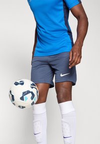Sininen Nike urheilupaita pyöreällä kauluksella, tumman siniset sivuakkentit, harmaat shortsit valkoisella logolla, valkoiset sukat ja kuvioitu jalkapallo.