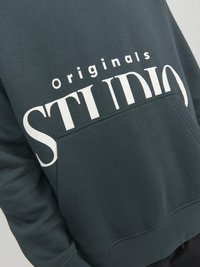 Mørkegrøn sweatshirt med trykt stort hvidt "STUDIO" og mindre "Originals" tekst. Har en lomme foran og blødt tekstureret stof.