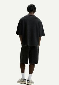 Černé oversize tričko a odpovídající šortky z texturované látky. Sportovní boty a bílé ponožky dotvářejí ležérní outfit.