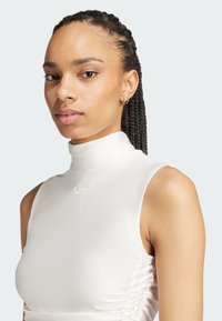 Witte mouwloze top met een hoge hals, voorzien van plissé aan de zijkant en een gebosseld logo op de borst. Gladde, elastische stof.