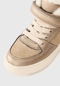 Metallisk gul sneaker med vita snören och en texturerad överdel. Har en vadderad krage och gummisula. Sömmen synlig längs sömmarna.