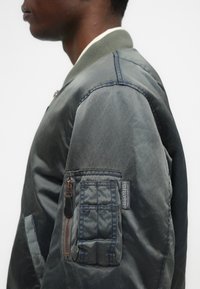 Jackets verde tipo bomber com uma textura suave e brilhante, gola canelada e um bolso com fecho no braço, apresentando um padrão acolchoado.