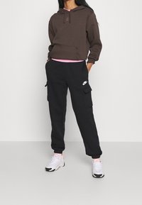 Hnědá mikina s kapucí a šňůrkou, černé cargo kalhoty s kapsami a bílé tenisky. Růžová košile viditelná pod mikinou.