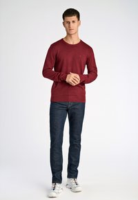 Bordowy sweter dzianinowy z okrągłym dekoltem, zestawiony z ciemnoniebieskimi dżinsami i białymi trampkami z kolorowymi akcentami. Prosty fason, bez wzorów.