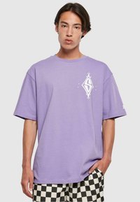 Purple short-sleeve t-shirt made of cotton, featuring a white diamond logo on the chest and a relaxed fit. Paired with black-and-white checkered shorts.