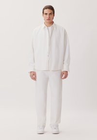 Camicia bianca con bottoni, dalla vestibilità comoda, abbinata a pantaloni bianchi abbinati e sneakers bianche, su uno sfondo neutro.