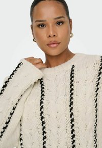 Pull en tricot torsadé crème avec des rayures horizontales noires, col rond et manches exagérées, présenté avec une finition douce et texturée.