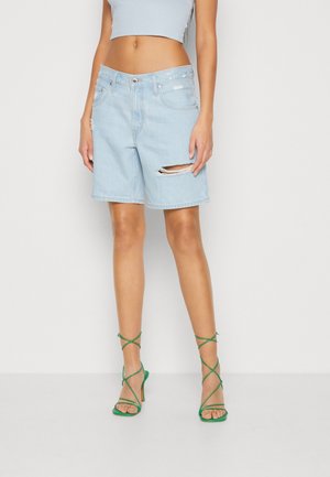 Femme portant un short en denim bleu clair déchiré jusqu'aux genoux, un top court et des sandales à talons hauts vertes à lanières, debout devant un fond clair uni.
