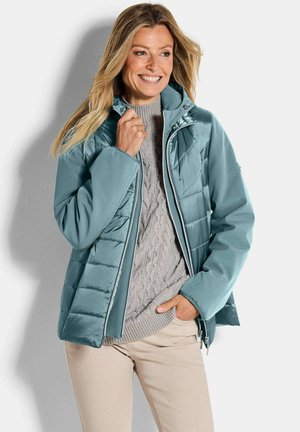 Femme souriante portant une veste matelassée bleu sarcelle par-dessus un pull tricoté gris à torsades et un pantalon beige, posant avec une main dans la poche.