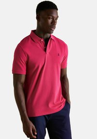 Muž v jasně růžovém polo tričku s krátkým rukávem a tmavě námořnických kalhotách, stojící s jednou rukou v kapse a dívající se do strany.