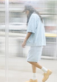 Svijetloplavi traper komplet s opuštenom košuljom i šortsevima, uparen s smeškim cipelama. Pozadina prikazuje vertikalne prozirne panele, stvarajući prozračan efekt.