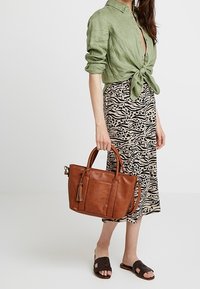 Brun skuldertaske i læder med kvastdetalje, rektangulær form og to håndtag. Bruges sammen med en grøn hørskjorte og et dyreprintet skørt.