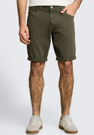 Homme portant un short vert olive retroussé, des baskets blanches et une chemise blanche, debout avec une main dans la poche devant un fond uni.