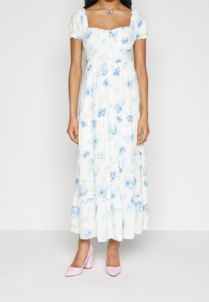 Woman wearing white maxi dress with blue floral print, puff sleeves, square neckline, pink block heel shoes, and a butterfly necklace.
