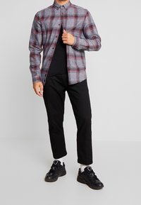 Camicia in flanella a quadri rossi e grigi con colletto button-down, dotata di maniche lunghe e orlo dritto, indossata sopra una maglietta nera e pantaloni neri.
