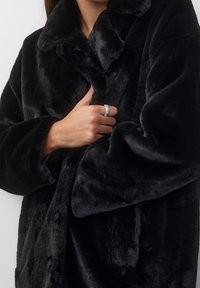 Cappotto in pelliccia sintetica nera con una texture soffice, colletto con risvolto e chiusura davanti. Una mano con un anello d'argento è stretta sul petto.