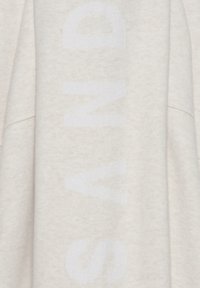 Hellbeige Sweatshirt aus strukturiertem Stoff. Mit großen, verblassten weißen Buchstaben auf der Vorderseite. Lockerer Schnitt und überschnittene Schultern.