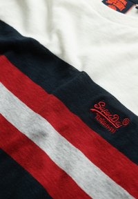 Camiseta a rayas en blanco, azul marino, rojo y gris claro. Presenta un logo bordado de "SuperDry" en rojo. Hecha de un suave y texturizado material de algodón.