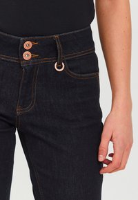 Jeans de denim oscuro con botones de cobre, costuras naranjas y una cintura ajustada. Presenta bolsillos frontales y una textura suave.