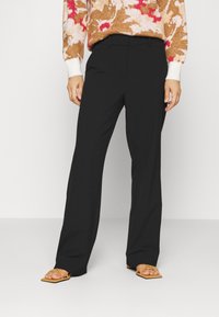 Pantaloni neri su misura con design a gamba dritta, caratterizzati da una texture liscia e vita alta, abbinati a sandali beige minimalisti.