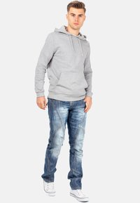 Un sweat à capuche gris avec cordons, associé à un jean bleu déchiré mettant en valeur des coutures diagonales, et des baskets blanches avec des accents rouges.