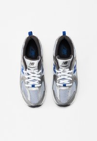 New Balance skriešanas apavi ar baltu acs augšu, pelēkiem akcentiem, ziliem detalizējumiem un baltām šņorēm. Mīkstināta zole ērtībai.