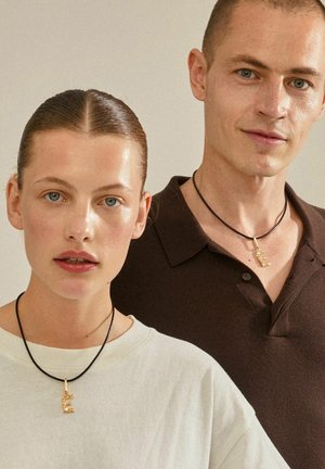 Gold initial pendants on black cords worn by a woman in a cream top and a man in a dark brown polo shirt, both with neutral backgrounds.