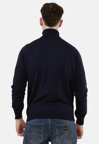 Maglione a maglia blu navy con colletto alto e maniche lunghe. Design aderente con polsini e orlo a coste. Indossato con jeans azzurri.